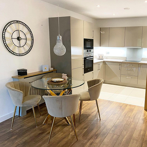 light dining and kitchen area, with warm wooden flooring, grey cabinets in kitchen, round glass table with gold legs and 4 beige velvet dining chairs. 