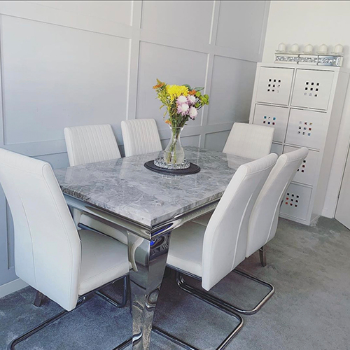 instagram image showing modern dinign area - 1 feature wall of pale grey wood panelling, pale grey carpeted floor and marble-effect table with chrome legs, with 6 white leather chairs. 