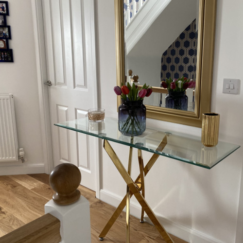 Upper landing at the top of a flight of stairs. Warm wooden floor and bannister. White walls. Gold chrome and glass console table sits at top of stiars, with vase of flowers. Large gold-framed mirror hangs above it, reflecting blue and gold geometric wallpaper over stairs. 