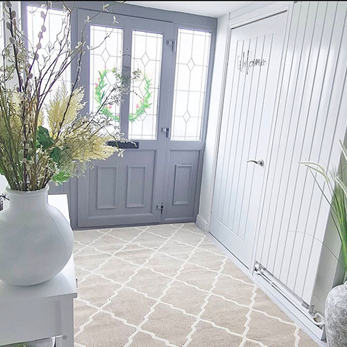 Bright entrance hallway with panelled grey front door and lots of windows. White walls and tiled floow with beige morrocan tile style rug on floor, and white wooden console table sporting white vase and grasses/dried flowers. Ceramic urn and leafy plant just visibile to right of frame.