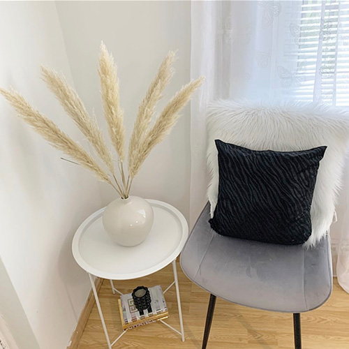Bright corner beside window. White metal side table with pampas grass in white vase. Grey velvet chair with black tapered legs and white shagyy cushion placed in corner