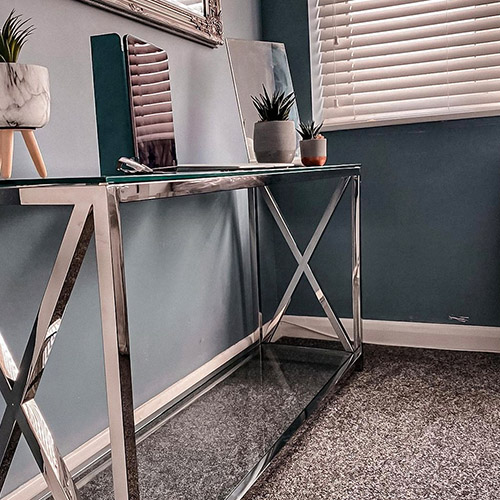 Corner of room / hallways with navy walls and grey carper. Silver chrome and glass console table in corner is being used as narrow desk, with laptop and pot plants on it. 