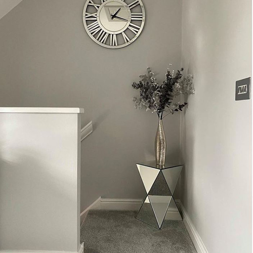 Upper landing at the top of a flight of stairs. Mirrored geometric side table sits in the corner, with vase/flowers on top. Grey carpet. One grey wall, one white wall.
