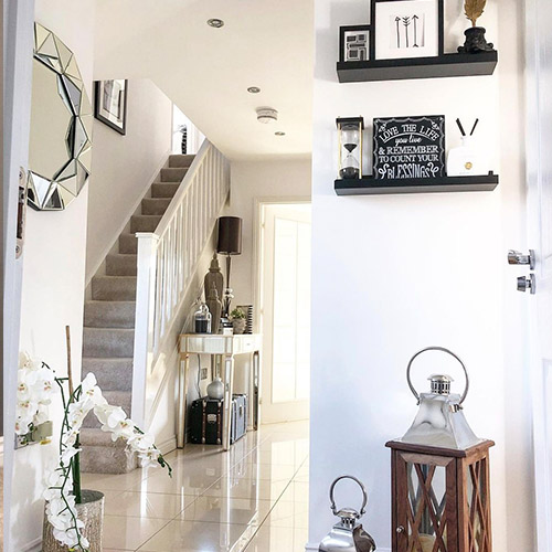 modern bright hallway with tiled flooring, mirrored console table against stairs banisters, round wall mirror, floating black shelving, matching accessories and potted orchid on floor.