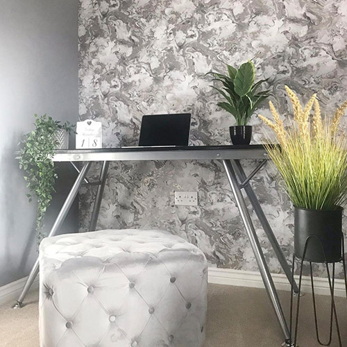 Corner of modern hallway featuring patterned grey wallpaper to one wall and grey plain wall to the other. Narrow desk with grey velvet cube cushion seat and lots of potted plants are placed in the corner.