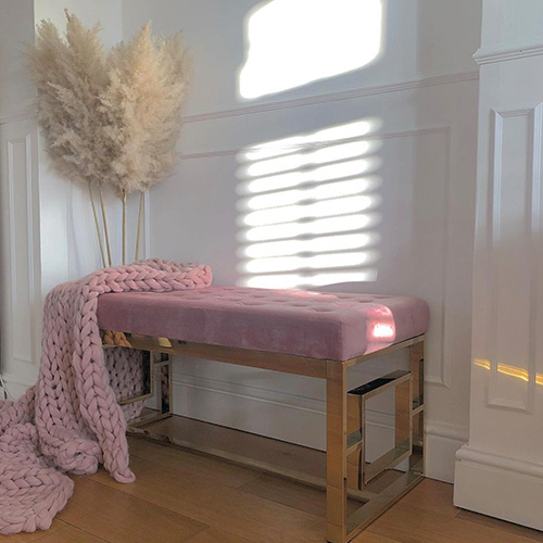 White hallway with wood panelling and warm yellow wood flooring. Art deco style bench seat placed in alcove, with gold chrome leg frame and pink velvet cushion seat. Accessorised with pink chunky knit blanket throw and pampas grass. Sun streams across the back wall. 