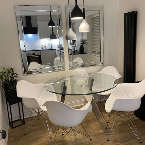 modern dinng room, warm wood floor and white walls, 2 large rectangular mirros on the wall, black pendant ligthing, round glass table with silver chrome legs, white chairs with chrome legs, black planter, white walls, black radiaote. Forsted glass globe ornament on table.
