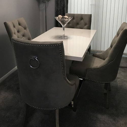 White glossy dining table with 4 velvet grey chairs with knockerback style. Grey carpet, grey wall, glass cnetrepiece filled with grey and white pebbles. 