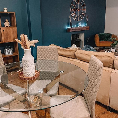 Bold petrol teal wall with large coc and fireplace, shared living space. Neutral shades in sofa and dining chairs, with accessories in natural tones - pampas grass in small eatehrn jug on table with gold legs. Sofa in beige velvet. Two velvet thros in teal and green. 