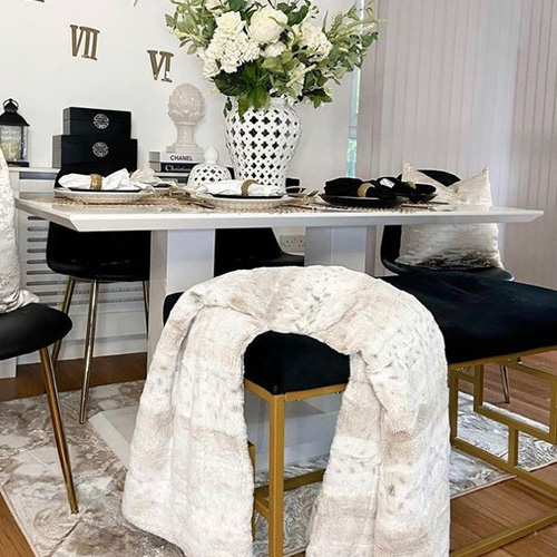Modenr dining room in black and white tones with pops of gold. White gloss table, with black faux leather chairs with gold legs. Black velvet bench seat with gold deco geometric leg frame. Table is dressed with black and white napkins in gold napkin rings, gold cutlery, white croket, white vase with white roses, gold wall clock and black items on far wall shelf. White throws and cushions on seats. 