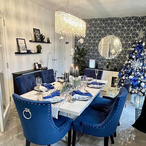 Bright dining room dressed for christmas. Far wall has blue and silver geometric wallpaper. Table is white gloss, seats are blue velvet with knockerbakcs. Round wall mirror, large crysal modern chandelier fixing, white tield floor, Christmas tree in blues and silevers. Black shelving on white walls