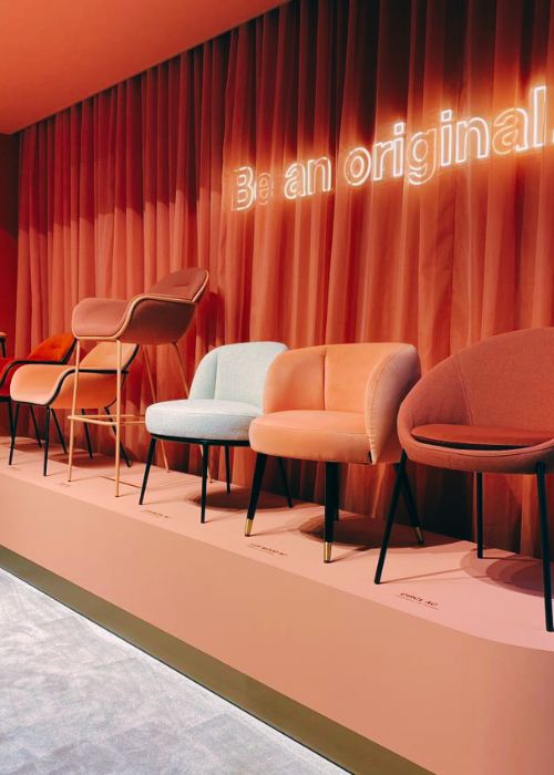 Stage with row of accents chairs in orange, yellow and white velvets and boucle fabrics, with black legs, against orange curtain with lighting that reads 'be an original'.