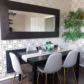 Modern dining room wit black gloss dining table and 6 faux leather grey dining chairs. Patterened wallpaper (gold geometric patterns) and green foliage. A wide rectangular mirror in a thick dark wooden frame is mounted horizontally on the wall above the table. 