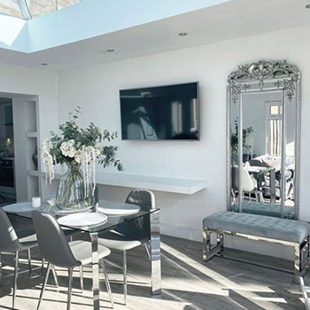 A bright and sunny room with glass ceiling, shows a grey tiled floor, a glass and chrme dining table with 4 grey leather seats, a long white floating shelf in  the far wall, and a grey velvet bench seat. A long vertical mirror hangs above the bench, in a dramatic silver and chrome frame.