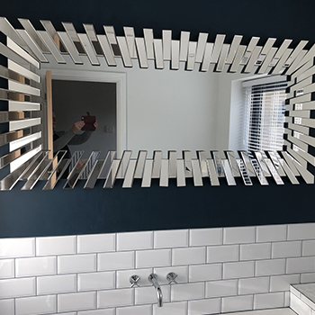 Bathroom mirror ideas - A bathroom wall above a bath  - white tiling in small rectangular horizontal brick pattern. Above this, a dark navy paint. A large rectangular mirror in starburst frame is hung horizontally on the dark paint. 