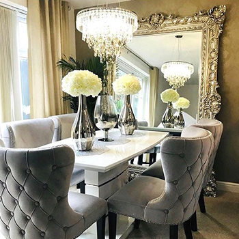 Dining room with dramatic gold emobssed wallpaper, white gloss dining chir, grey velvet sears with flower arrangement. A modern glass drop chandelier hangs above, and a very large mirror is set against the back wall, in a dramatic gold gilt frame. 