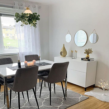 Simple dining room with neutral walls, wooden floor and grey rug beneath a white gloss dining table with chrome legs, and 4 grey velvet chairs. a minimalist chest of drawers is against the far wall, with simple accessories. 3 mirrors hang above it - all round, in simple gold thing band frames. Central mirror is larger than the other 2. 