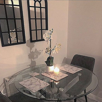 A shaded dining area in a corner of a room. 2 window mirrors hang on the wall behind the table, in dark wooden frames with arched tops.