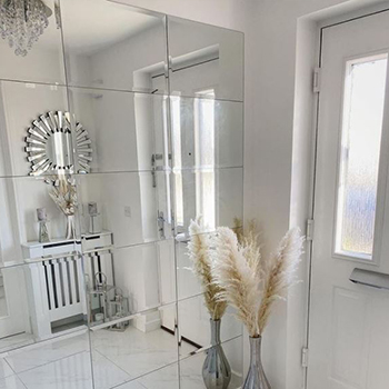 Entryway mirror ideas. Narrow hallways. One wall is entirely covered in plain mirror tiles. We see the opposite wall reflected in this, which shows a simple white wooden console table and a round mirror in mirrored starburst frame in its reflection. A dried flower arrangement is of grasses is also shown. 