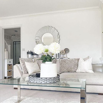 Bright living room with pale neutral shades of furnishings. Glass and chrome coffee table in foreground with flower arrangement. A large round mirror hangs on the back wall, in a large chrome/mirrored sunburst frame. 