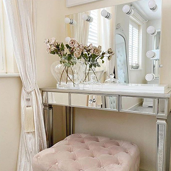 Bright bedroom with mirrored console table and a large rectangular vanity mirror with large round lightbults. A vase of roses is placed on the vanity console table. A pink plush velvet square stool sits in front of it. 