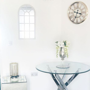 DIning room mirror ideas - a white room with a round glass dining table that has chrome legs. a silver clock hangs on the wall to the right, and a white-framed window mirror with arched top hangs on the left wall. 