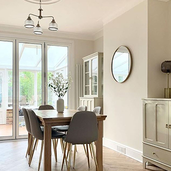 A rustic style kitchen with wooden table, grey faux leather dining chairs and wooden floor. Wooden dresser and cabinet can also be seen, painted. A chimney breast with no fireplace in the far wall, with a round mirror in a simple thin frame hanging on it.