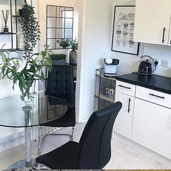 Modern small-ish kitchen. Decor is white and black theme, with round glass dining table, 2 black leather chairs, white cupboards with black countertops and black door handles. Lots of greenery and plants. A rectangular mirror hangs on the far wall, reflecting the plants. 