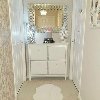 Narrow hallway decorated in beige and cream. A white wooden dresser is placed at the far end, with a lamp and accessories. A rectangular mirror with frame comprising of overlapping mirrored circles is hung above it, reflecting the lamplight.