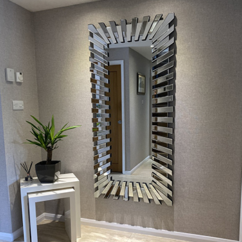 Hallway mirror ideas - a corner of a hallway. A set of white stacked nested tables in the corner with a potplant. on the facing wall. The floor is pale laminate, the walls are grey wallpaper. A large rectangular mirror in a starburst frame is hung on one wall.