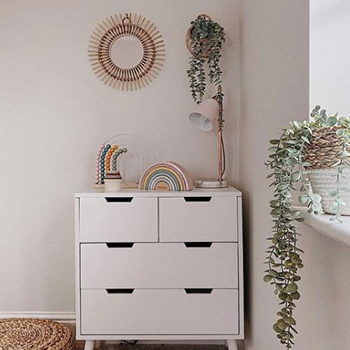 White room, showing a white wooden chest of drawers, with a simple pink and brass lamp, and wooden rainbow children's toys. A plant is on the way in a wicker holder, beside a small-ish round mirror in a gold sunburst frame. 
