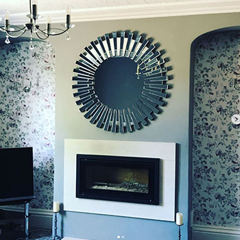 Living room mirror ideas. Round mirror in a starbust frame hung over a fireplace, with statement floral wallpaper in the alcoves beside fireplace. A simple chandelier syle light fixing also hangs int the shot. 