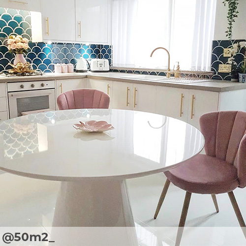 Instgram image from @502m_ featuring modern kitchen with dining table. Kitchen is white and glossy with white floor tiling, white counters with gold accents, teal fish scale style tiles. In foregorund, white high gloss mushroom table wth 2 pink velvet dining chairs. Pink and gold accessories to decorate.  
