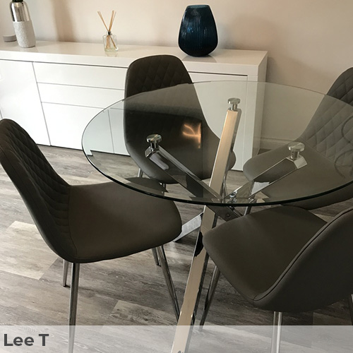 Dark wood floor with cool tone. Furniturebox UK Selina round glass dining table with chrome legs and 4 black Corona dining chairs with black legs. White simole minimalist sidebaord in background with white and black accessories. 