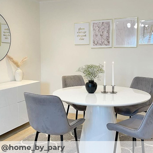 IG image from @home_by_pary. Bright modern dining area with white walls and sideboard, simple round mirror and white/pink/gold artwork on wall. Warm tone light wood effect flooring and grey morrocan tile rug beneath white high gloos mushroom table and 4 grey velvet chairs. 