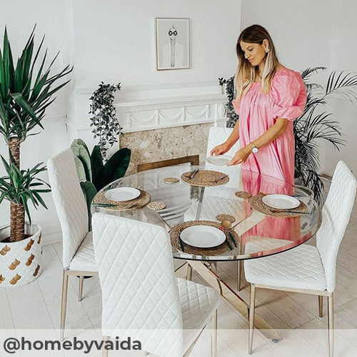 instagram image from @homebyvaida featuring furniturebox uk novara gold round glass dining table with 4 white mialn faux leather chairs with gold legs. Stylish dining area with white walls, feature fireplace pale wooden flooring and potted plants in white/gold pots. Vaida is setting the table, smiling and wearing a pink dress.