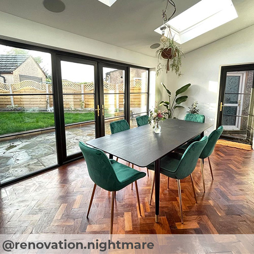 Instagram image from  @renovation.nightmare. Open dining space with large wall to wall french windows and black frames, and sky lights. Dark wood herringbone parquet flooring beneath modern black dining table with tapered legs and 6 green velvet dining chairs with gold legs. Pot plants hang from ceiling and stand in cornner. 