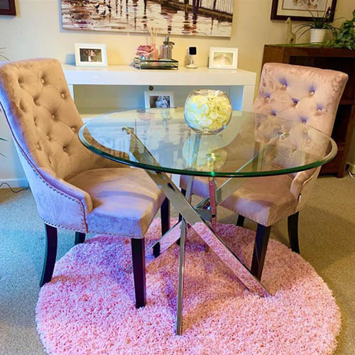 small round glass dining table with 2 grey velvet knockerback chairs on a pale grey carpet with a round pink rug beneath the table, giving a pinkish glos to table legs.