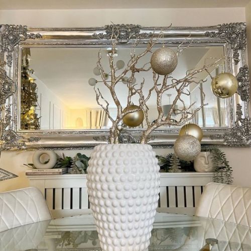 round glass dining table 2 whute velvet dining chairs, whitevase with gold branches and gold baubles, large ornate silver mirror in background.