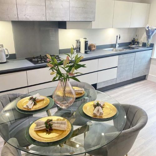 round glass dining table with gold starburst legs, 4 grey velvet seats, gold charger plats and napkins, vase of lillies.