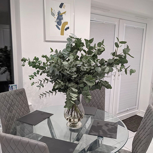 round glass table with chrome starbusrt legs, 4 grey faux leather chairs with silver legs, and clear glass vase filled with sage-green greenery