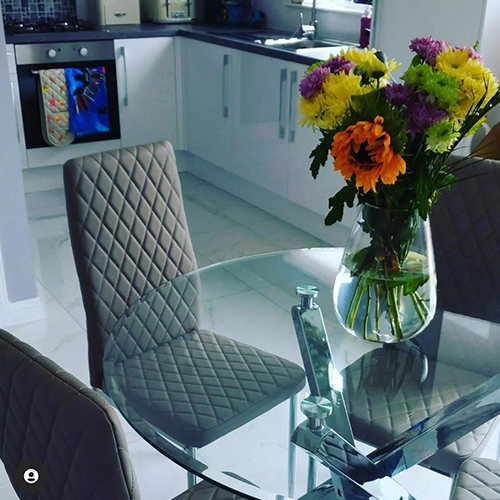 round glass table with chrome starbusrt legs, 4 grey faux leather chairs with silver legs, and clear glass vase filled with colourful fresh flowers