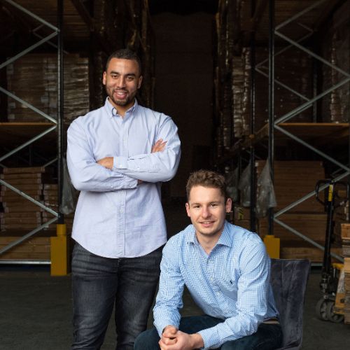 funriturebox founders monty george and dan beckles smile in warehouse. Dan, a black man, stands with arms crossed and large smile. Monty, a white man, is seated with hands clasped on knees, smiling