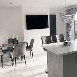 Modern open space kitchen diner with grey and white breakfast bar and 2 tall barstool seats, large wall mounted TV on chimney breast in rear, with round glass dining table in alcove to left of TV, with 4 grey velvet dining chairs. 