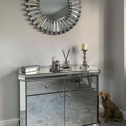 Alcove in Grey room featuring stylish mirrored console table with large round wall mirror above in a mirrored starburst frame, decorated with scent diffuser and glass candlestick. 