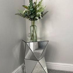 corner of room with grey walls and carpet with mirrored bedside table / plinth featuring triangular angled mirrored sections. Base of flowers placed on top.
