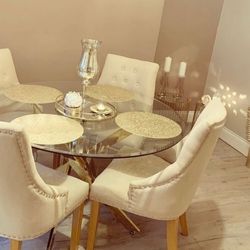 Warm beige and cream dining area, featuring round glass dining table with gold chrome starburst legs, 4 cream tall back chairs with wooden legs, and 4 gold-cream placemats on table with tall vase. 2 Short gold wire frame side tables placed in aclove in background, with 2 tall candlesticks and candleholder, casting reflections on wall. 