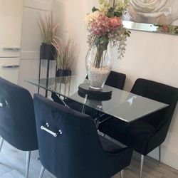 Bright dining area featuring rectangualr glass dining table and 4 black velvet tub chairs, with black centre mat and glass vase with white pebbles and pink/cream and green floral arrangement. Narrow alcove in rear with 2 black plant pots on tall legs, both filled with pink tinged pampass grasses. 