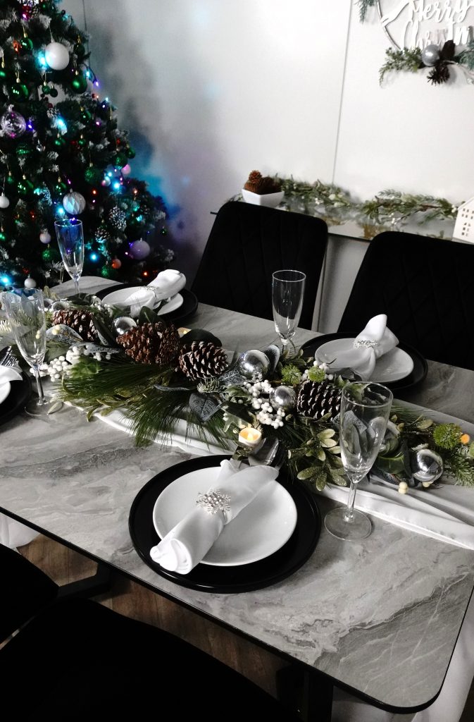 Carson melamine topped grey marble effect dining table with 4x black velvet dining chairs, with christmas tree in background and 2 wreaths laid back to back in centre of table