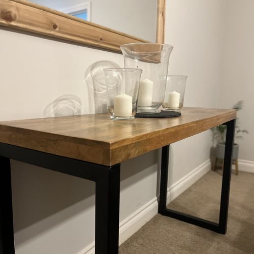 Wooden industrial style console table with solid wood top and black metal legs, three hurricane lamps with pillar candles, under large wall mirror.